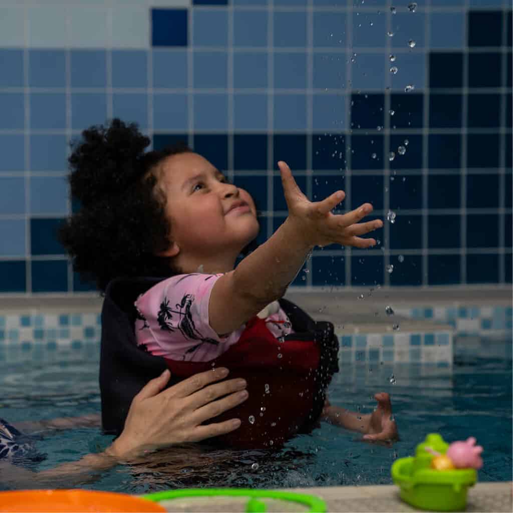 Little girl in hydrotherapy pool beams with outstretched hand to catch water sprinkling from above