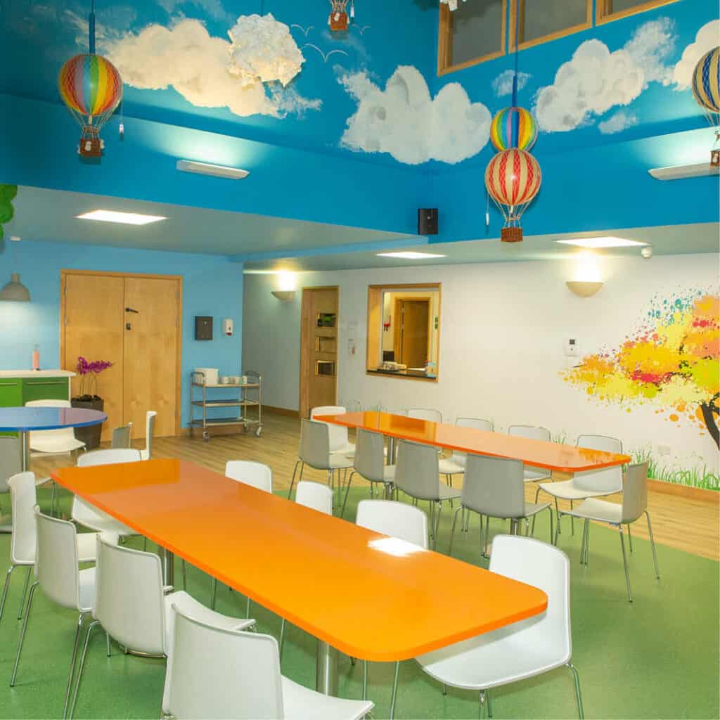 Bright colourful space with long orange dining table, white changes and colourful painted ceiling like the sky