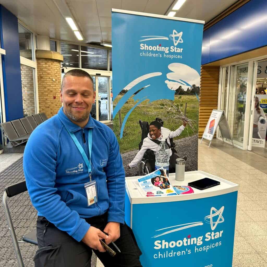 A man in a blue shooting star jumper, wearing an id badge, sat next to a promotional stand with shooting star children's hospices branding.