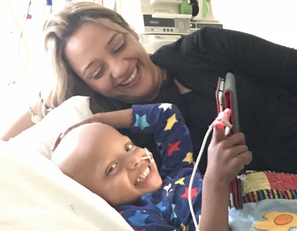 a boy in a hospital bed looking at the camera, with his mum lying next to him looking down at him.