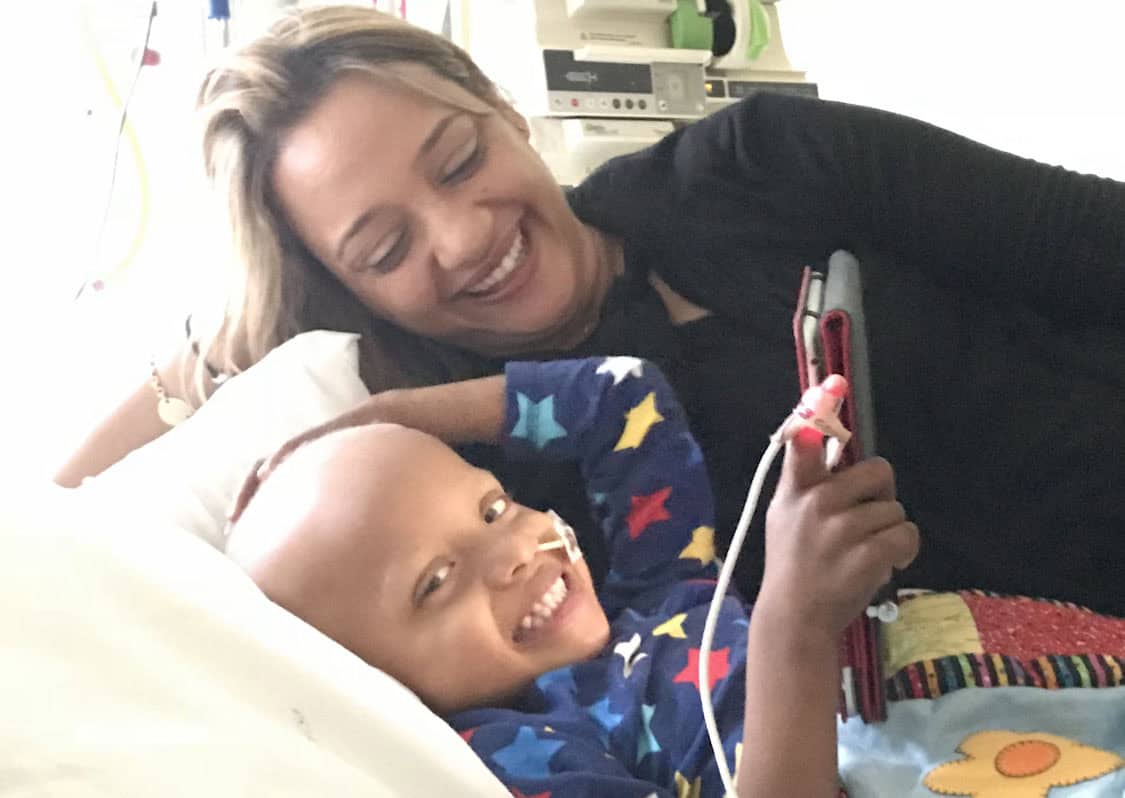 a boy in a hospital bed looking at the camera, with his mum lying next to him looking down at him.