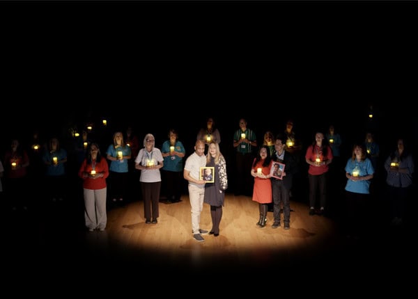 A couple standing on a stage holding a photo of their son, surrounded by Shooting Star staff holding candles.