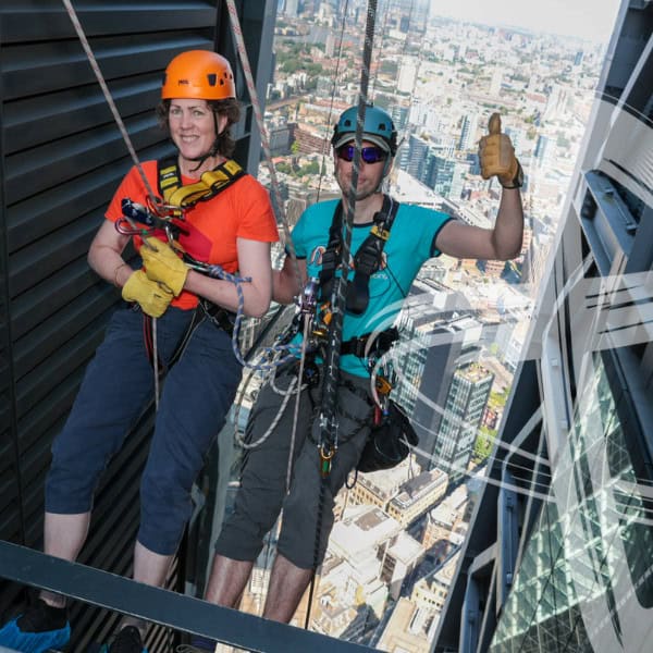 A nervous looking woman in an abseiling harness being guided over the top of a tall building