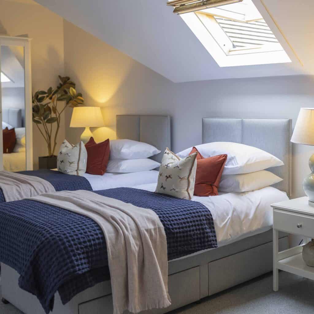 A bright bedroom with two twin beds made up with white sheets, layered blue and grey throws, and orange and patterned grey cushions. Two bedside tables with lamps sit between the beds, with a plant in the corner, a white mirror on the wall and a vaulted ceiling window bringing in natural light.